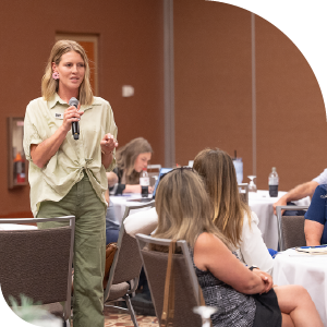 Woman holds a microphone and speaks to a full room of peers at a workforce event