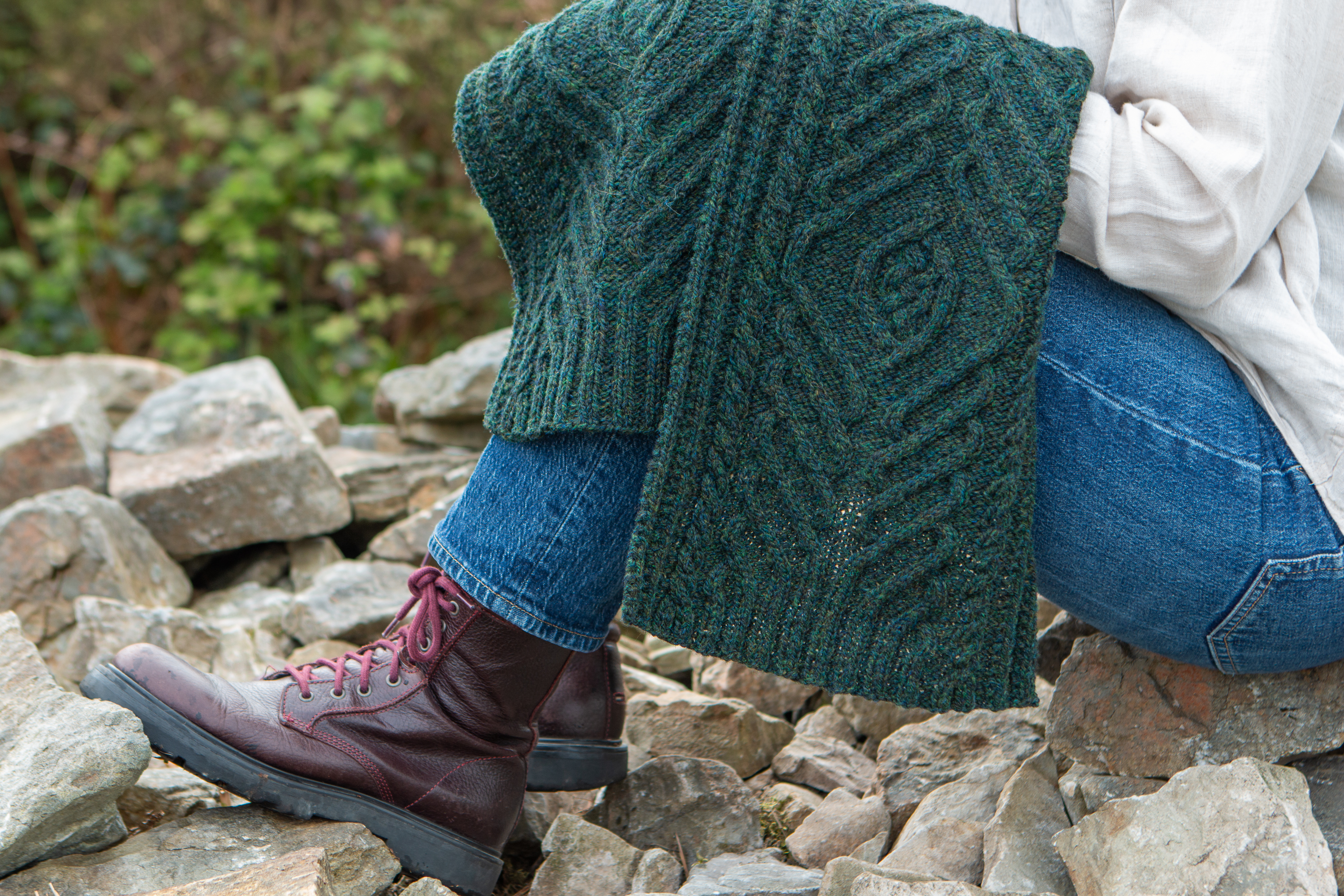 A hand-knit dark green cabled scarf being held over a persons blue jeans and doc martins.