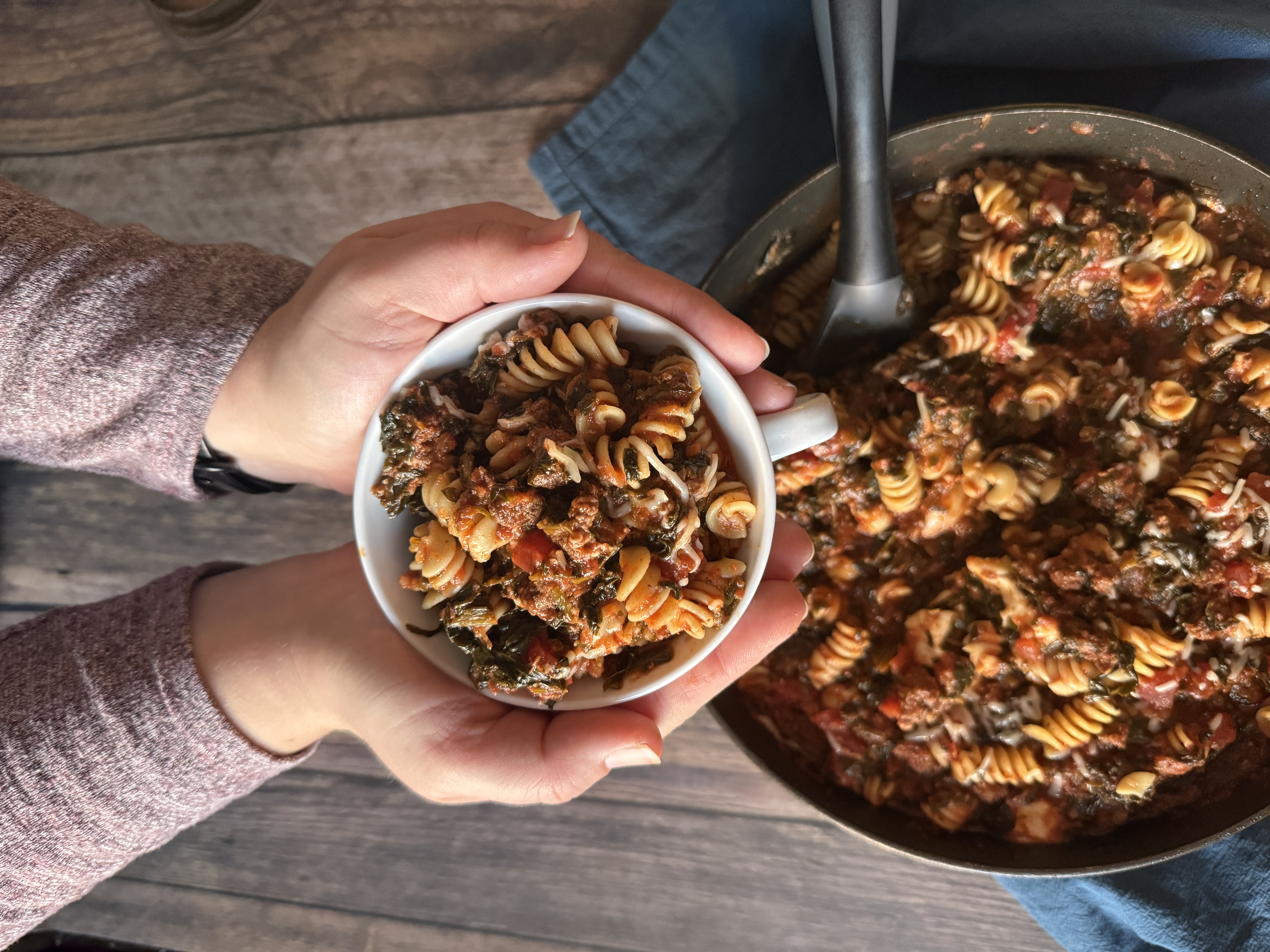 bowl of pasta in hands over pan of pasta with black and grey spoon over wood table with blue tablecloth