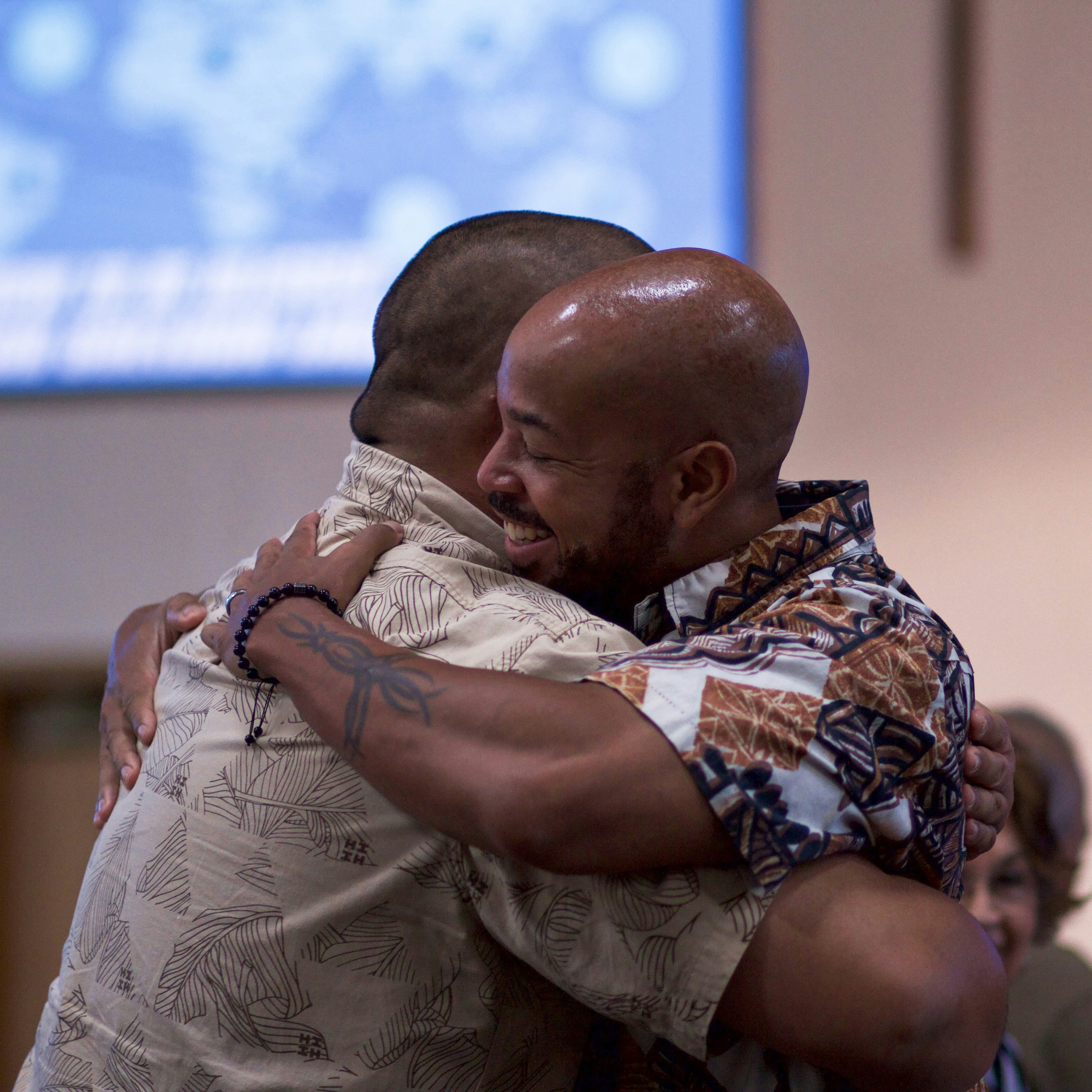 Two men embrace at an indoor gathering; one smiles as they hug.
