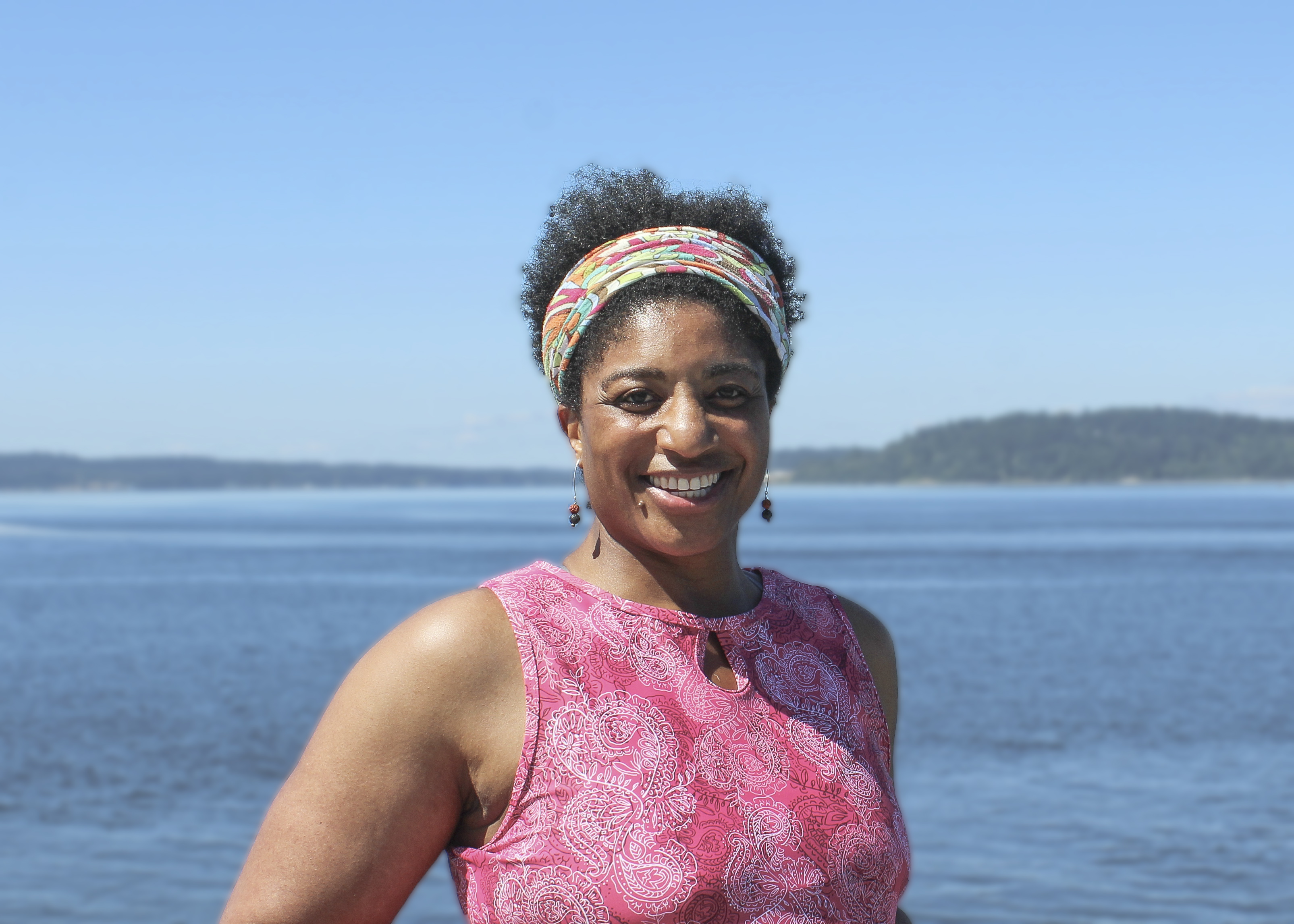 black woman wearing a headband and sleeveless, pink tunic. Standing in front of a body of blue water and blue sky, with land in the background. 