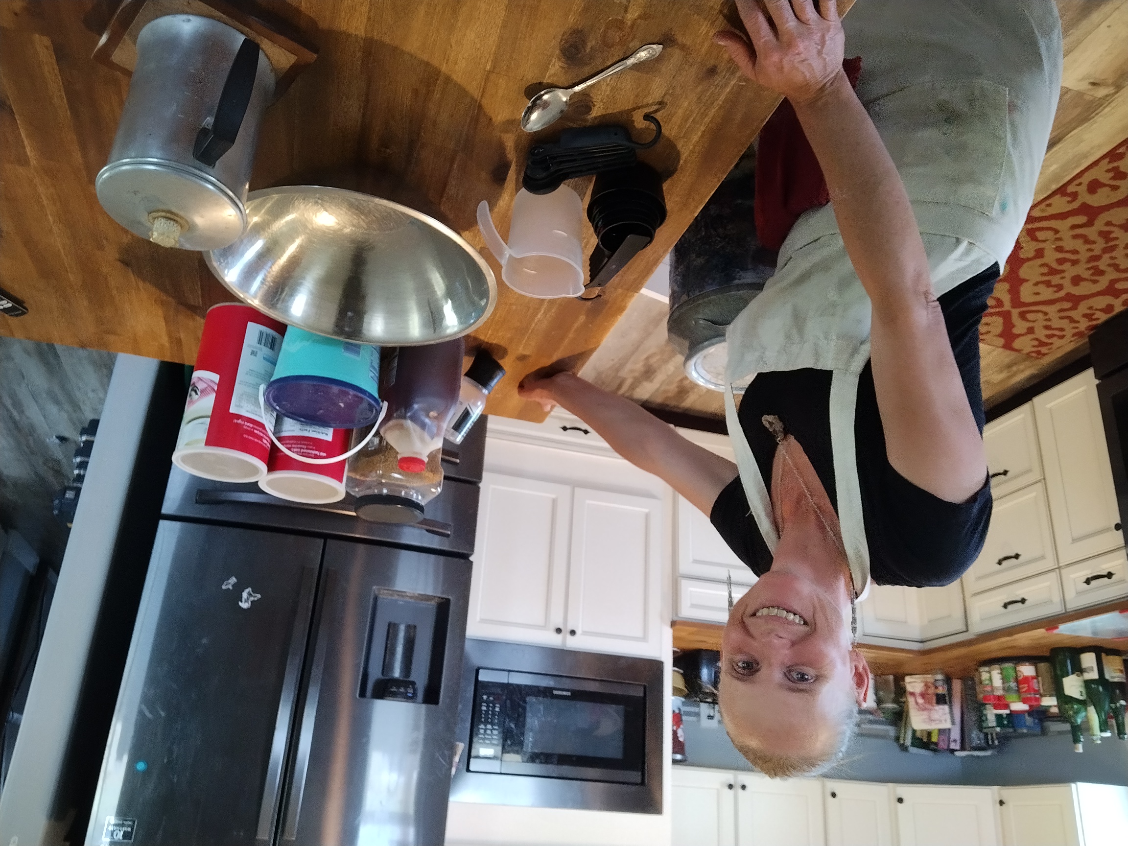 Smiling women, long hair, wearing jewelry and apron. Standing in kitchen behind wooden kitchen bar with baking supplies on it. Kitchen in background.of free standing oven/stovetop with arms crossed confidently