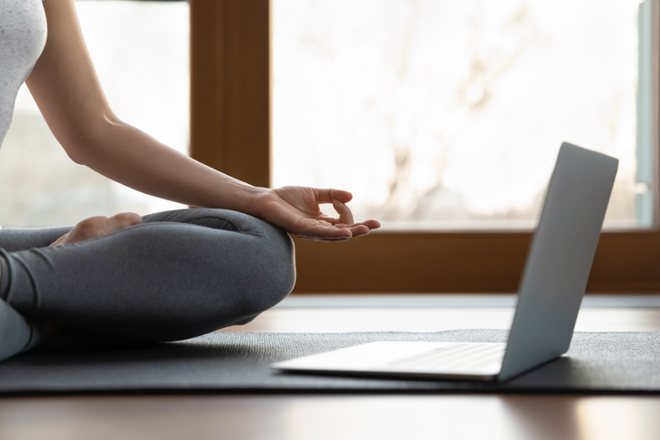 Silver laptop computer resting on a yoga mat with seated woman wearing gray tights and white top in front of a window
