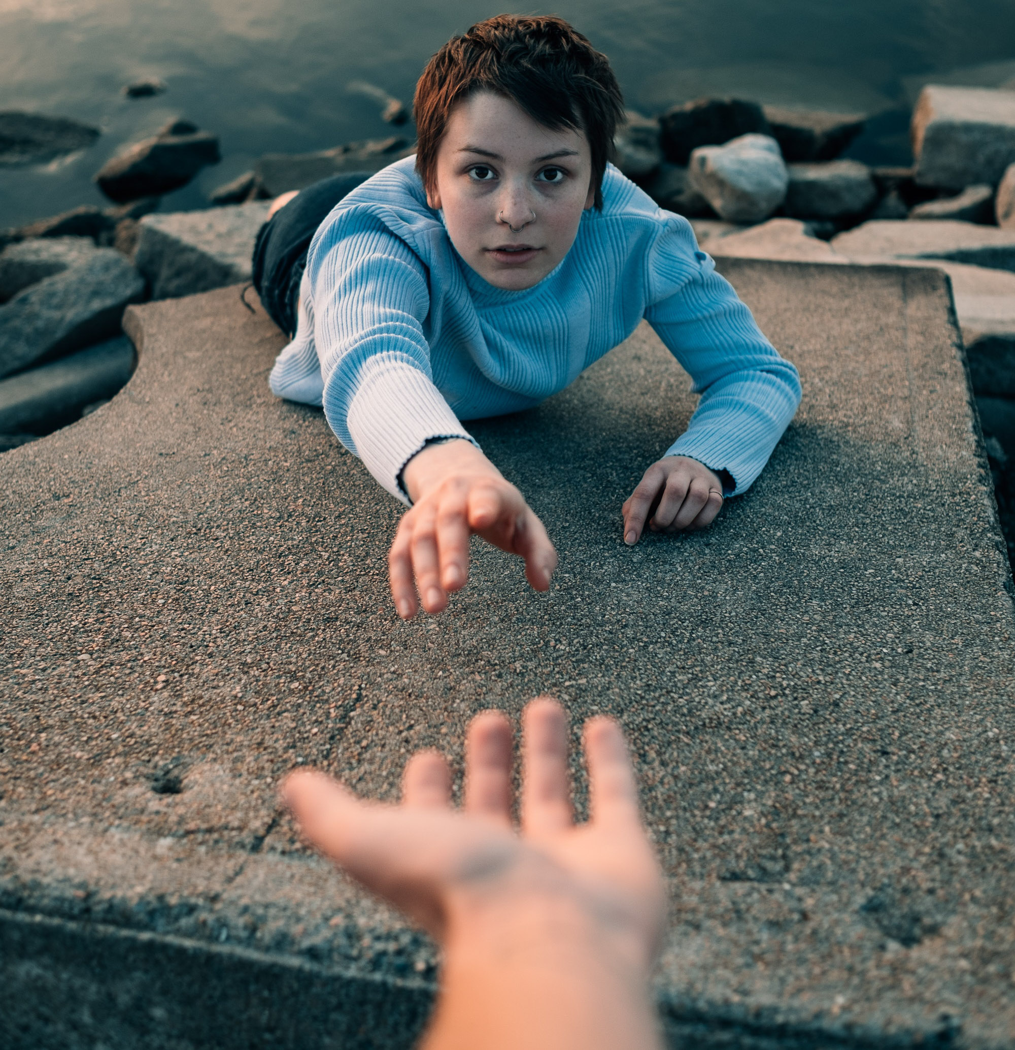 An individual is seen hanging above rocks and a body of water, reaching for another hand in the frame.