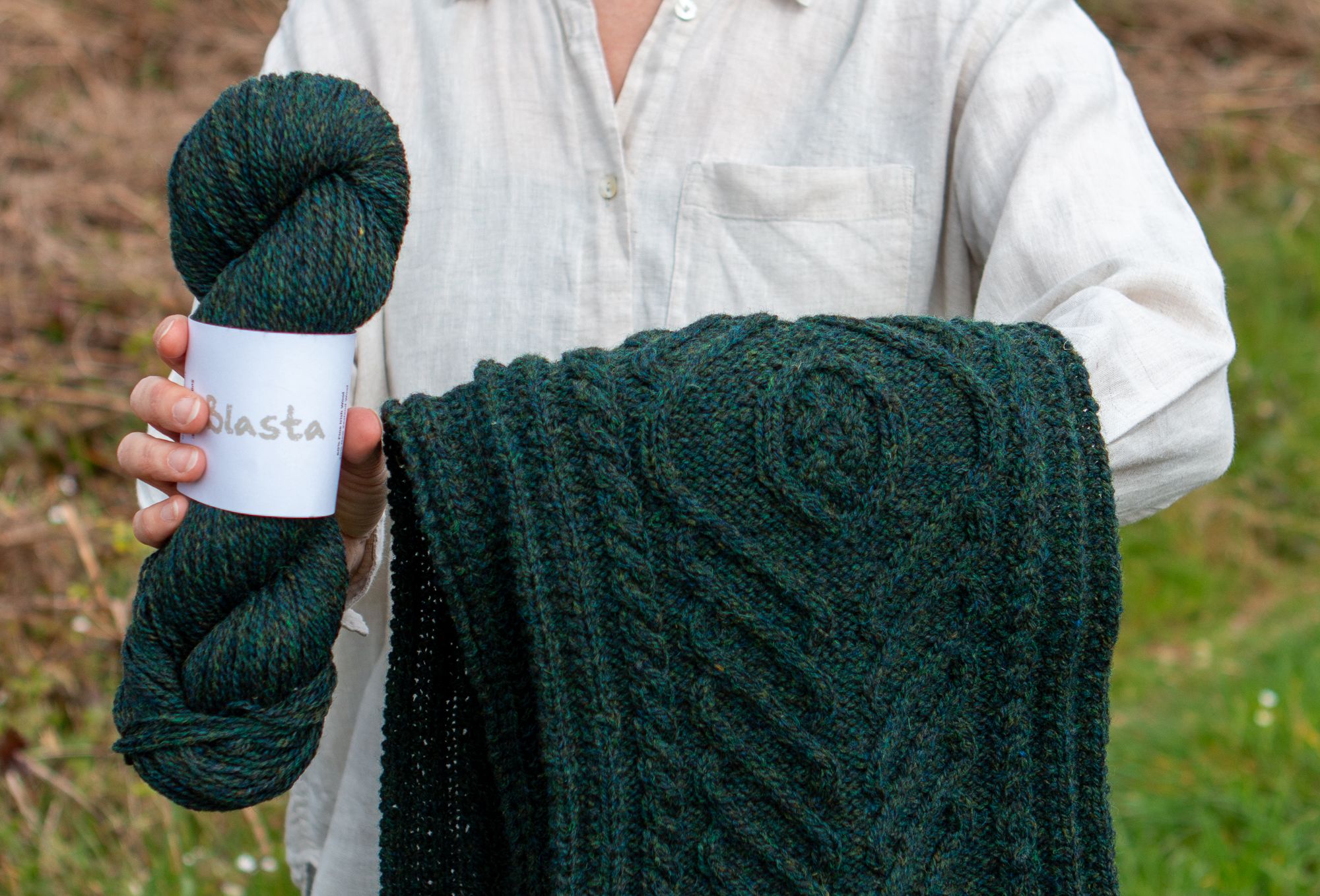 Person holding a skein of dark green yarn with a label reading Blasta and a hand-knit cabled scarf hanging over their other arm.
