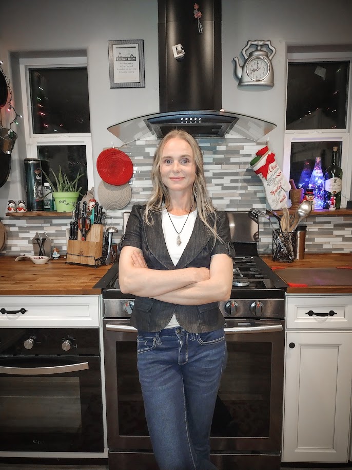 Smiling women, long hair, wearing jewelry and a business jacket with blue jeans. Standing in kitchen in front of free standing oven/stovetop with arms crossed confidently