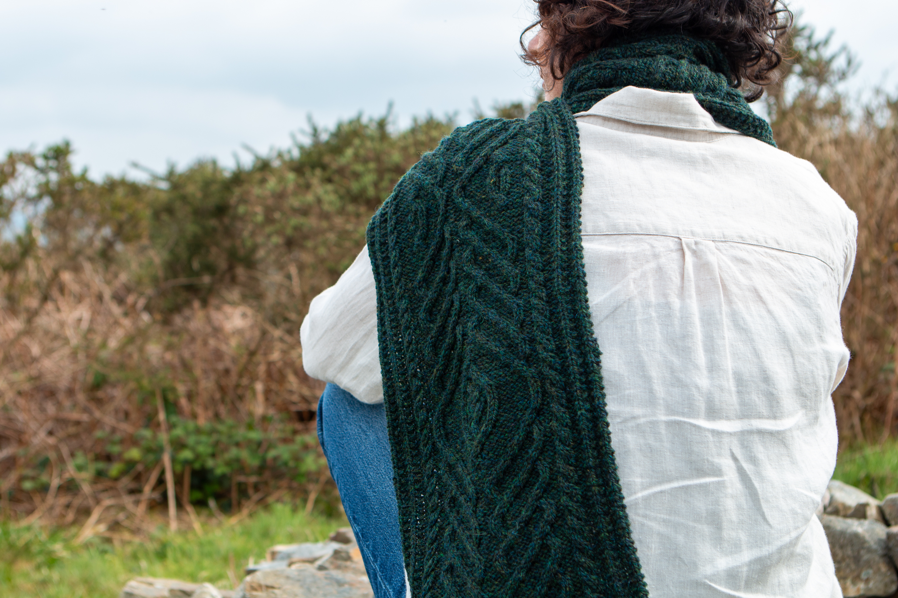 A hand-knit cabled scarf lying over the shoulder of a person sat down.